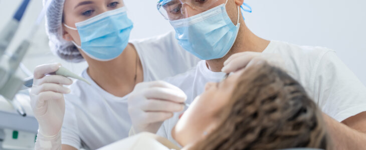 Low angle shot of a male dentist and his assistant examining a female patient's teeth. Young girl laying calm on the dental chair with mouth open wide.
